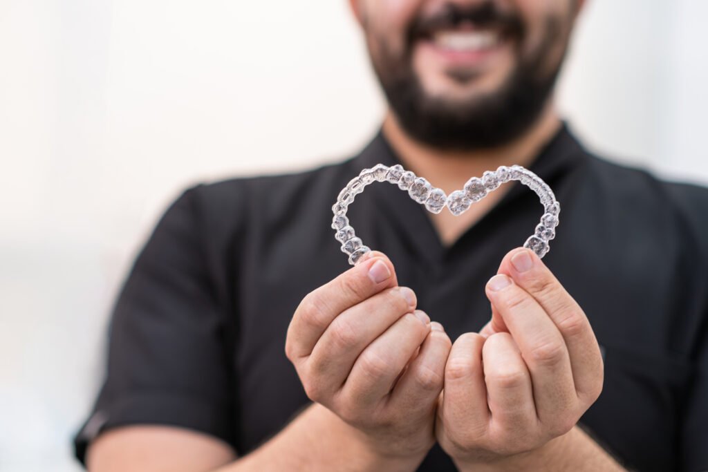 Smiling doctor holding heart made of clear aligners for professional teeth correction in clinic