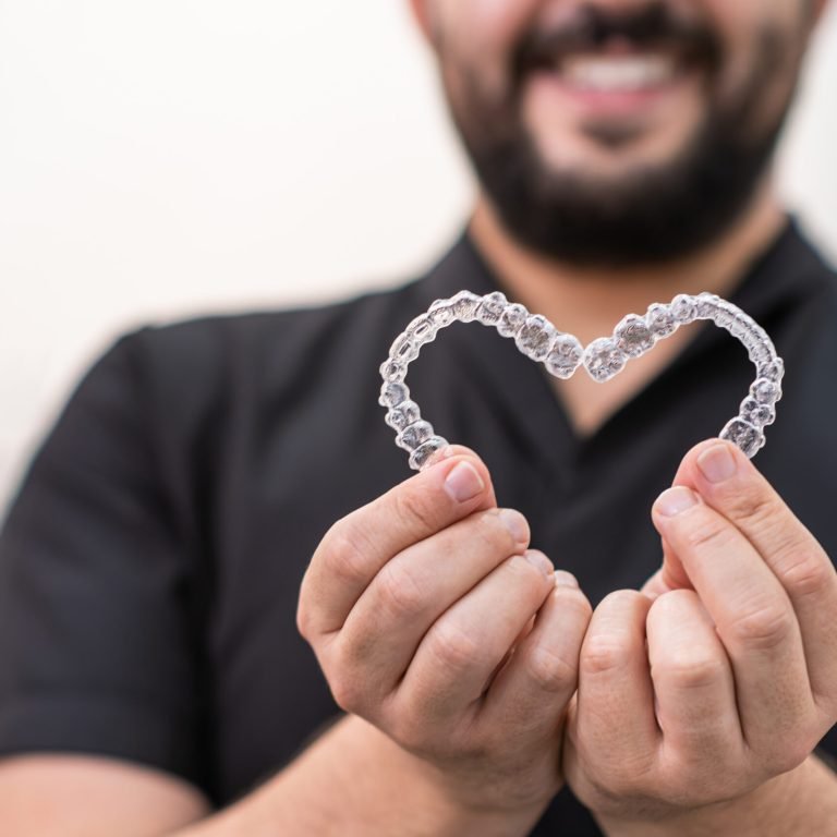 Smiling doctor holding heart made of clear aligners for professional teeth correction in clinic
