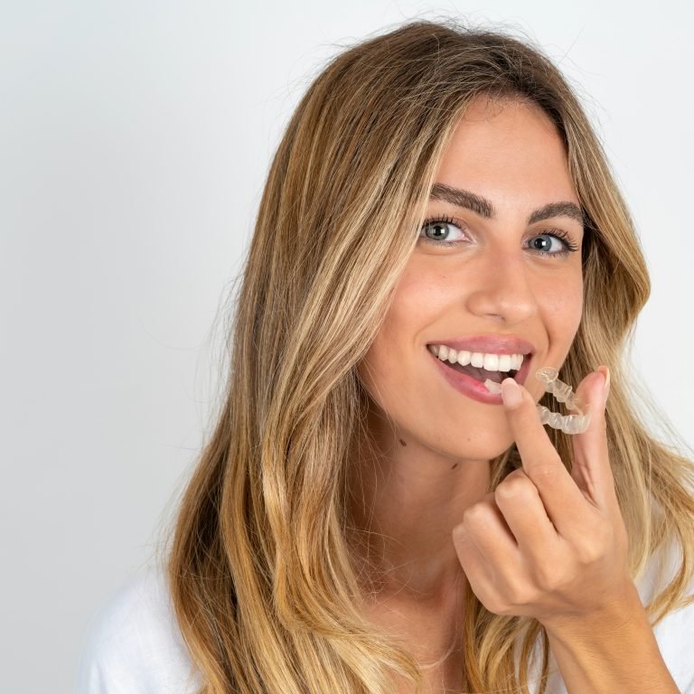 young blonde woman wearing white shirt over white studio background holding an invisible aligner ready to use it. Dental healthcare and confidence concept.